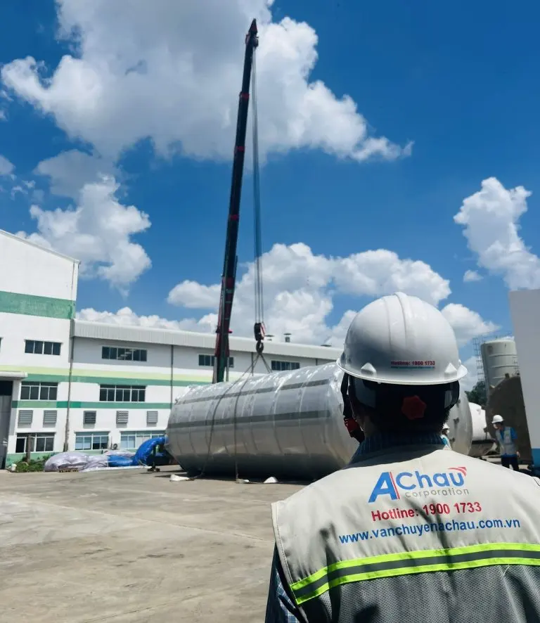 Nhân viên Á Châu mặc áo phản quang in hotline 1900 1733 giám sát cẩu bồn inox công nghiệp lên xe tại nhà máy