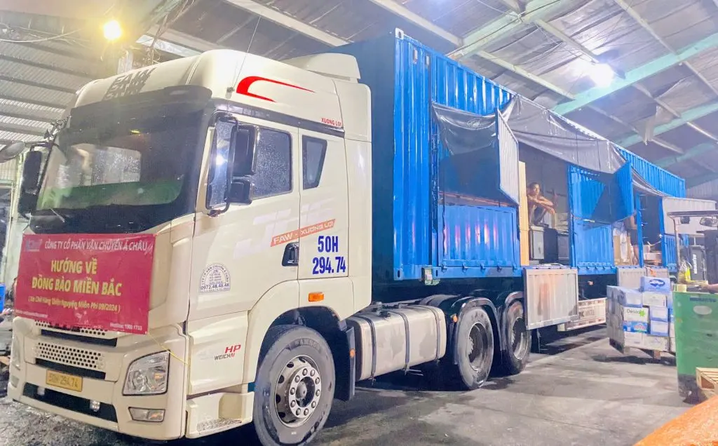 Đầu kéo container biển số nền vàng của Á Châu đang ăn hàng trong kho, treo băng rôn chuyến xe chở hàng cứu trợ đồng bào miền Bắc
